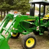 Zielony traktor John Deere 2130 z ładowaczem czołowym stoi na podwórku, gotowy do pracy.