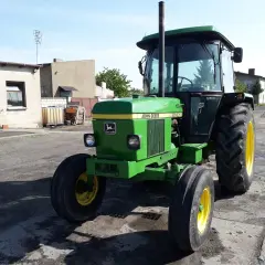Zielony John Deere 2140 z żółtymi felgami stoi na podwórku. W tle budynki gospodarcze i pole.