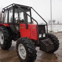 Czerwony ciągnik Belarus 952 z kabiną i osłoną, stojący na błotnistym podwórzu. W tle pole z resztkami śniegu.