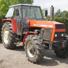 Pomarańczowy Ursus 1224 z kabiną stoi na drodze gruntowej, otoczony zielenią drzew i krzewów.
