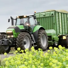Zielony ciągnik rolniczy z przyczepą pełną zielonki na tle pola i żółtych kwiatów.