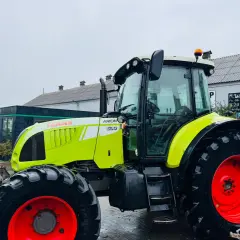 Jasnozielony ciągnik Claas Arion 630 z czerwonymi felgami stoi na mokrym placu przed budynkiem.