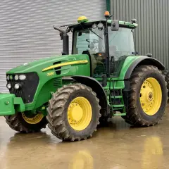 Zielony John Deere 7830 z żółtymi felgami stoi na mokrej posadzce przed budynkiem gospodarczym.