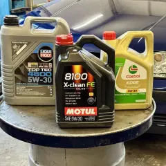 Trzy kanistry z olejem silnikowym: Liqui Moly, Motul i Castrol, na tle niebieskiego samochodu.