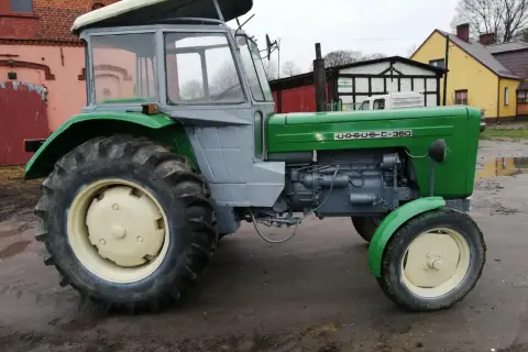 Zielony Ursus c355 z szarą kabiną, gotowy do pracy na błotnistym podwórku.