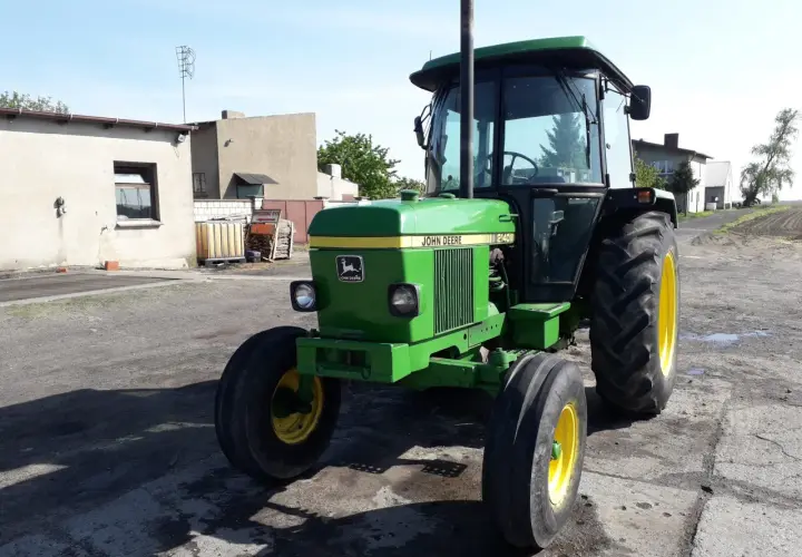 Zielony John Deere 2140 z żółtymi felgami stoi na podwórku. W tle budynki gospodarcze i pole.