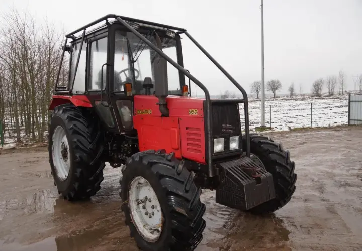 Czerwony ciągnik Belarus 952 z kabiną i osłoną, stojący na błotnistym podwórzu. W tle pole z resztkami śniegu.