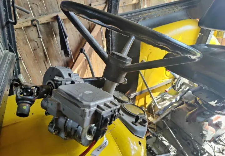 Mechanizm wspomagania kierownicy w żółtym ciągniku. Widoczna kolumna kierownicy, wał napędowy i elementy hydrauliczne.