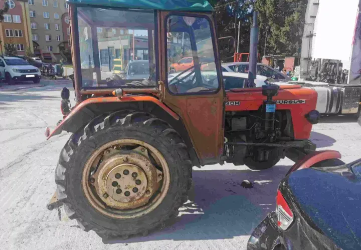 Pomarańczowy Ursus 30, jeden z najtańszych ciągników rolniczych używanych, z kabiną i dużymi kołami, stoi na placu obok samochodów.