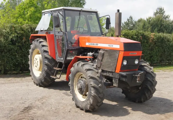 Pomarańczowy Ursus 1224 z kabiną stoi na drodze gruntowej, otoczony zielenią drzew i krzewów.