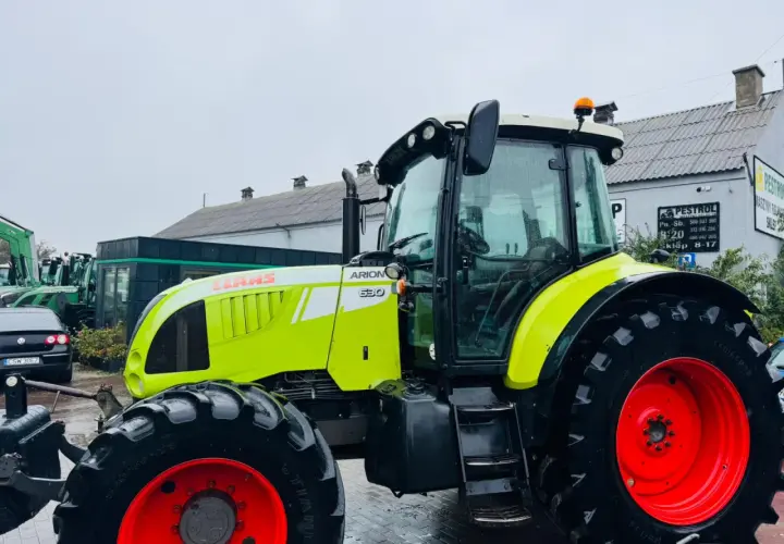 Jasnozielony ciągnik Claas Arion 630 z czerwonymi felgami stoi na mokrym placu przed budynkiem.