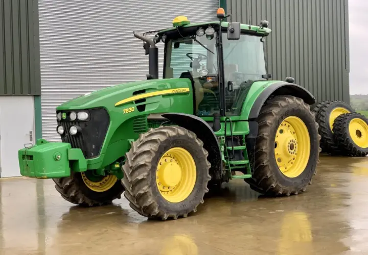 Zielony John Deere 7830 z żółtymi felgami stoi na mokrej posadzce przed budynkiem gospodarczym.