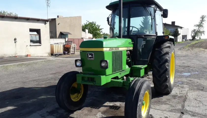 Zielony John Deere 2140 z żółtymi felgami stoi na podwórku. W tle budynki gospodarcze i pole.