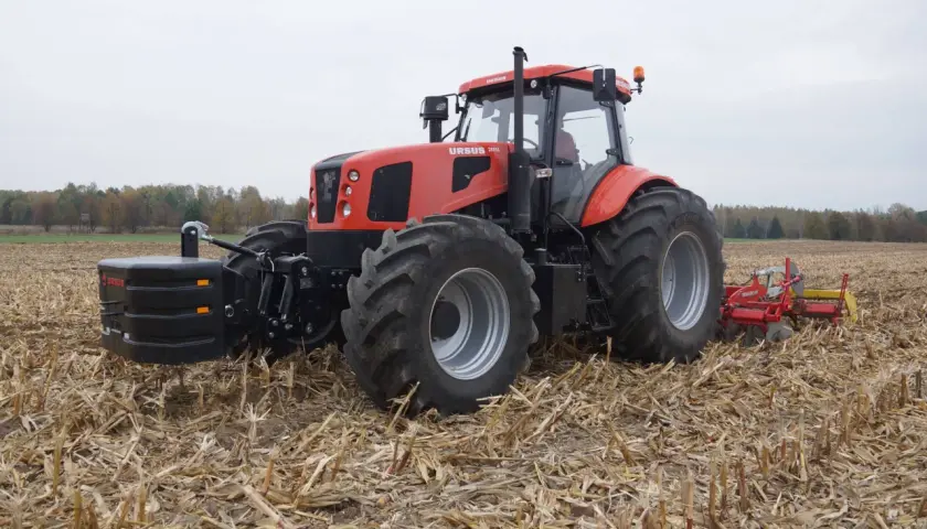 Czerwony Ursus 28014 z obciążnikiem z przodu i pługiem z tyłu pracuje na polu po żniwach.