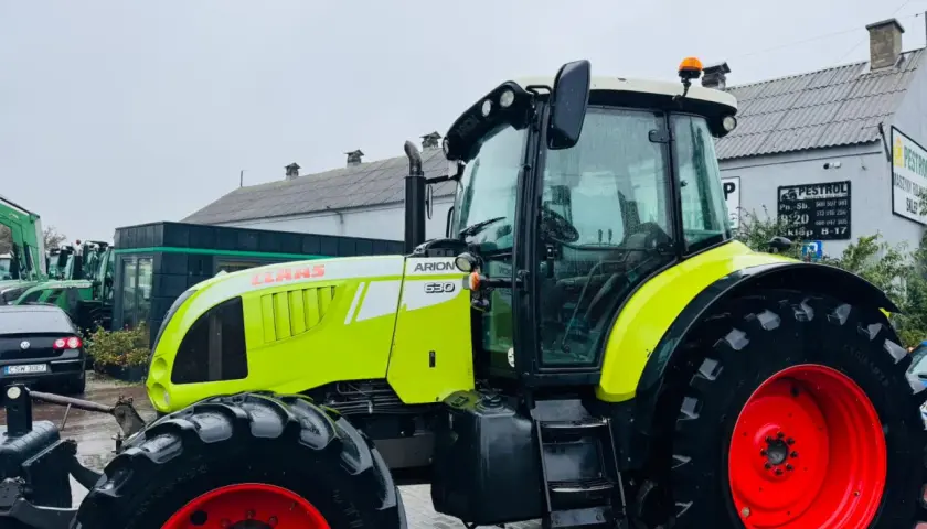 Jasnozielony ciągnik Claas Arion 630 z czerwonymi felgami stoi na mokrym placu przed budynkiem.