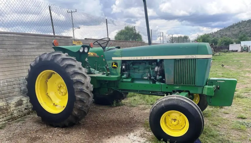 Zielony John Deere 3130 z żółtymi felgami stoi na żwirowym podwórku, gotowy do pracy.