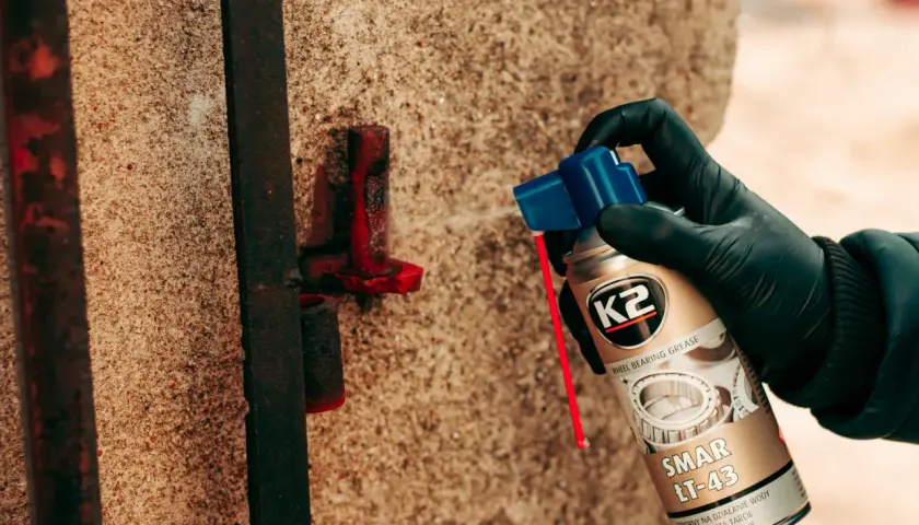 Ręka w czarnej rękawiczce aplikuje smar K2 LT-43 na zardzewiały element metalowy, chroniąc go przed korozją.
