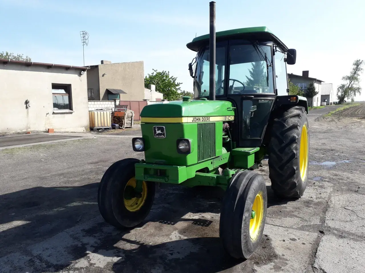 Zielony John Deere 2140 z żółtymi felgami stoi na podwórku. W tle budynki gospodarcze i pole.