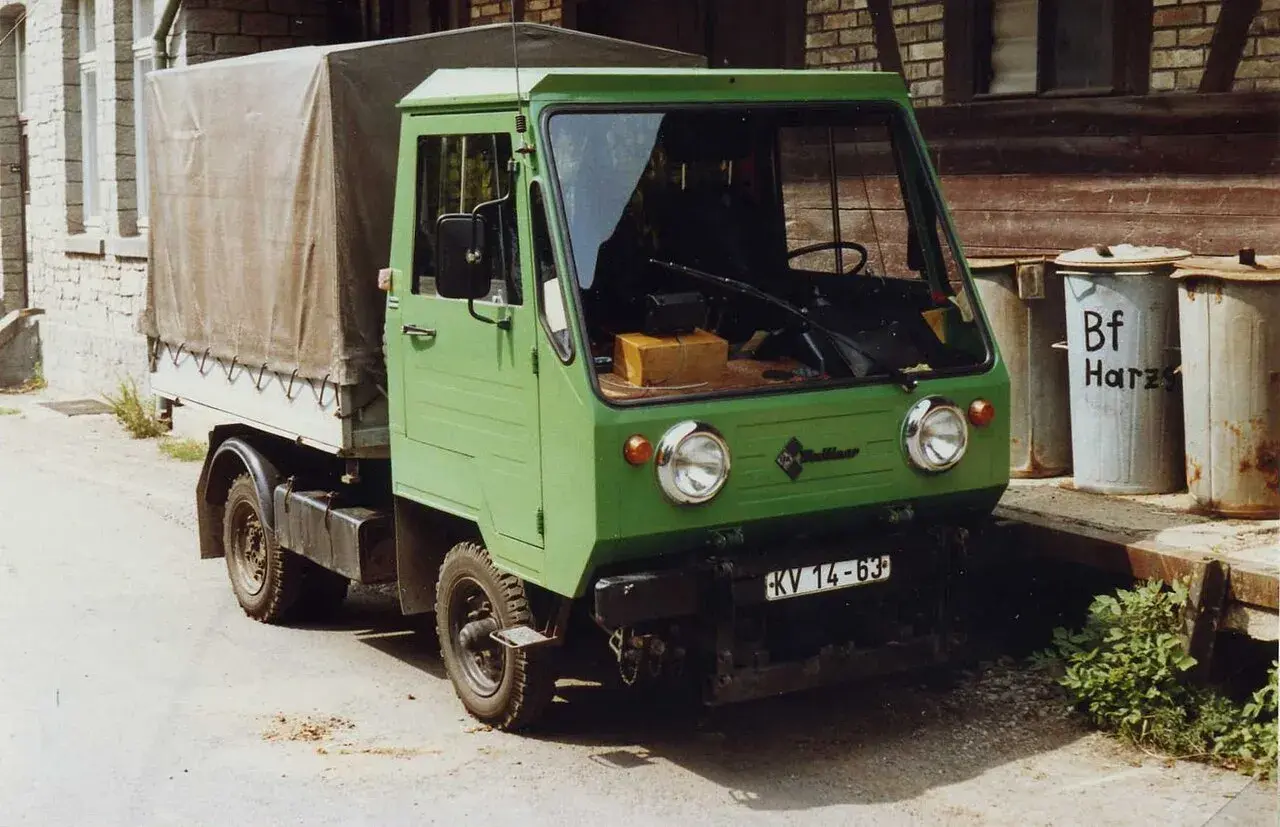 Zielony Multicar M25 z plandeką, gotowy do drogi. Obok śmietniki z napisem "Bf Harz".