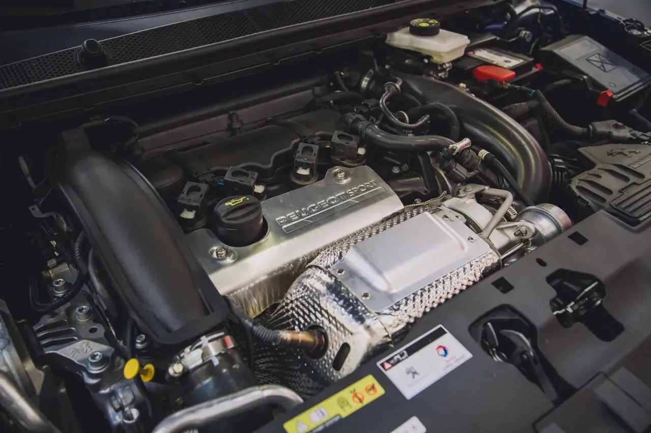 Silnik 1.6 PureTech z napisem "PEUGEOT SPORT" na metalowej osłonie. Widać turbosprężarkę i elementy układu dolotowego.