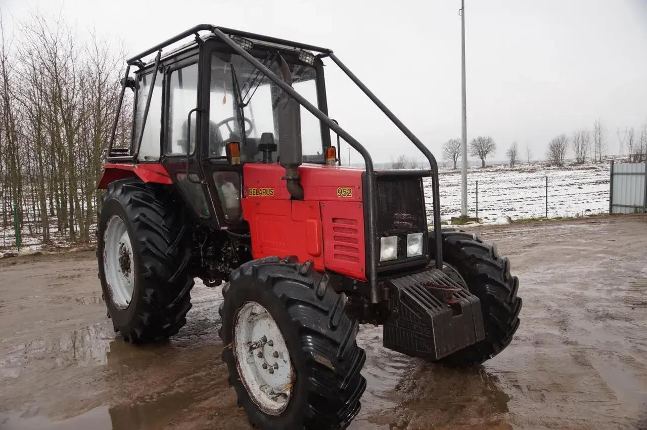 Czerwony ciągnik Belarus 952 z kabiną i osłoną, stojący na błotnistym podwórzu. W tle pole z resztkami śniegu.