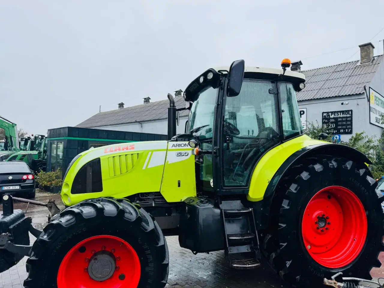 Jasnozielony ciągnik Claas Arion 630 z czerwonymi felgami stoi na mokrym placu przed budynkiem.