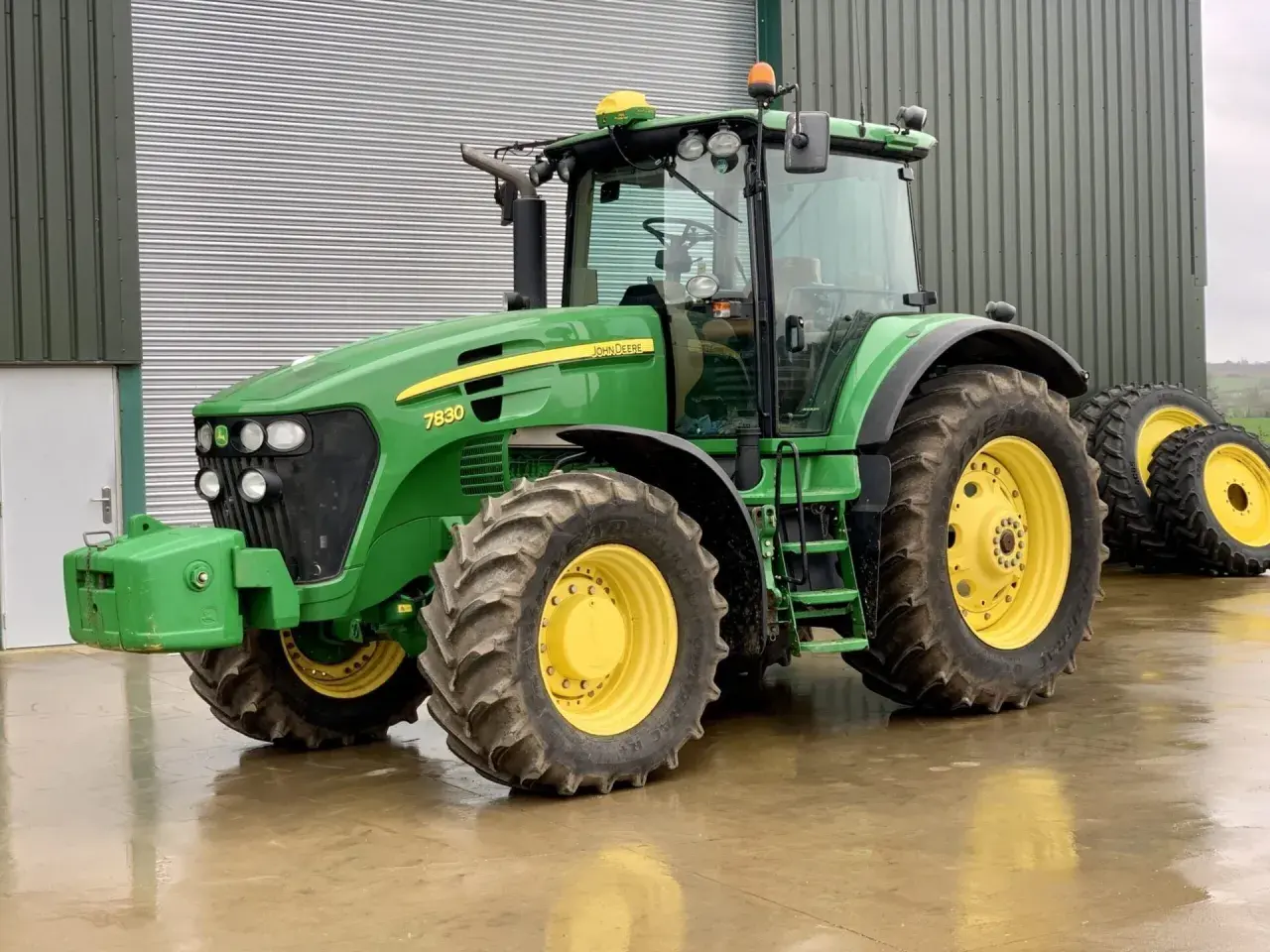 Zielony John Deere 7830 z żółtymi felgami stoi na mokrej posadzce przed budynkiem gospodarczym.