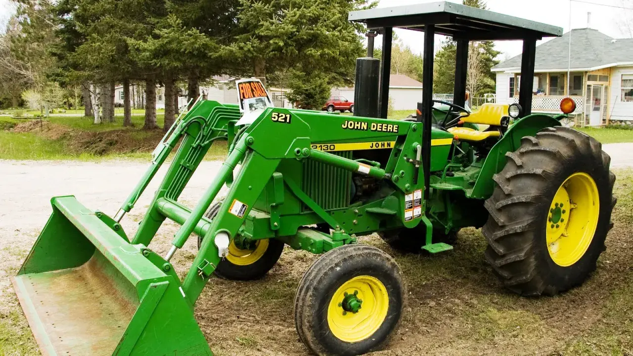 Zielony traktor John Deere 2130 z ładowaczem czołowym stoi na podwórku, gotowy do pracy.
