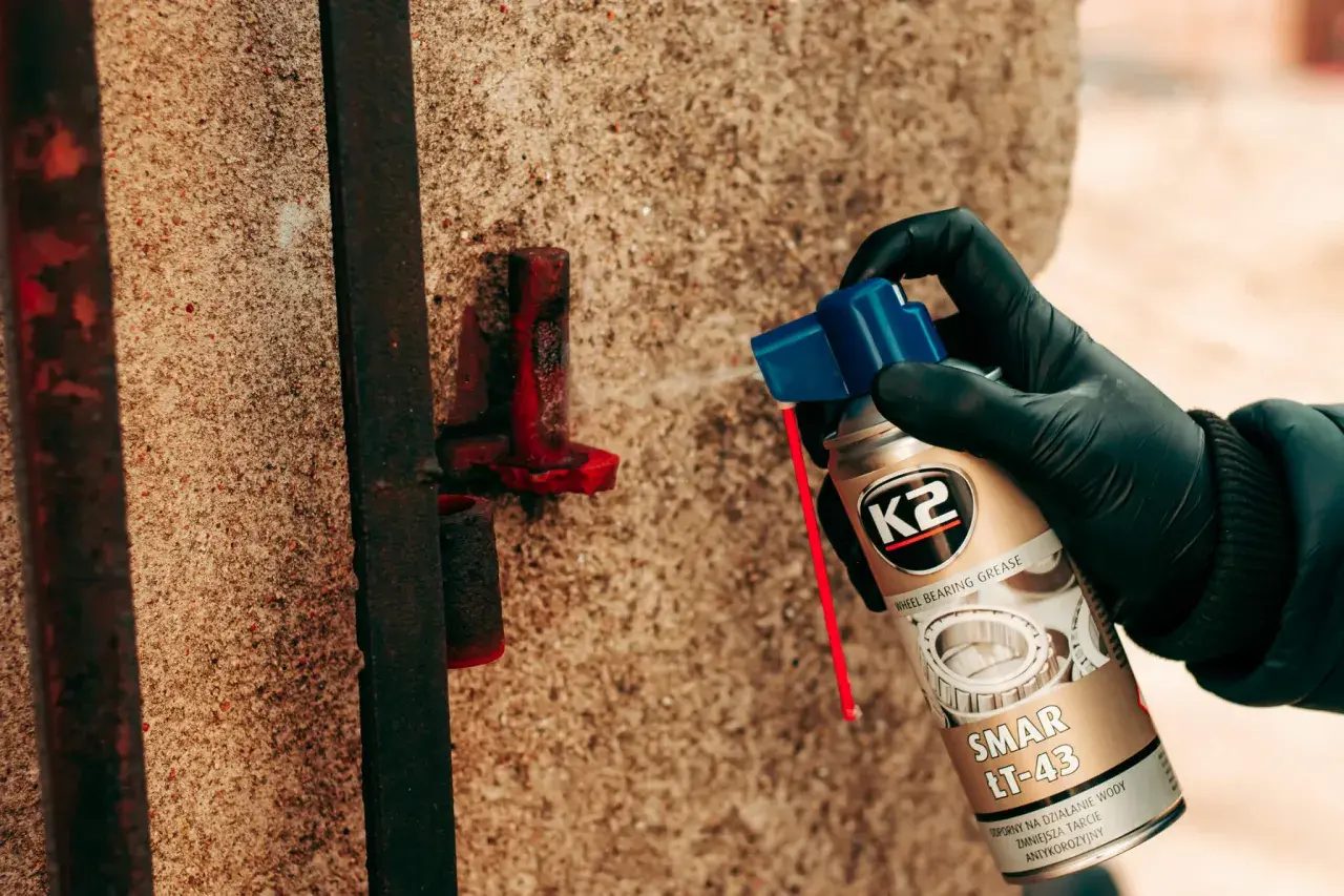 Ręka w czarnej rękawiczce aplikuje smar K2 LT-43 na zardzewiały element metalowy, chroniąc go przed korozją.