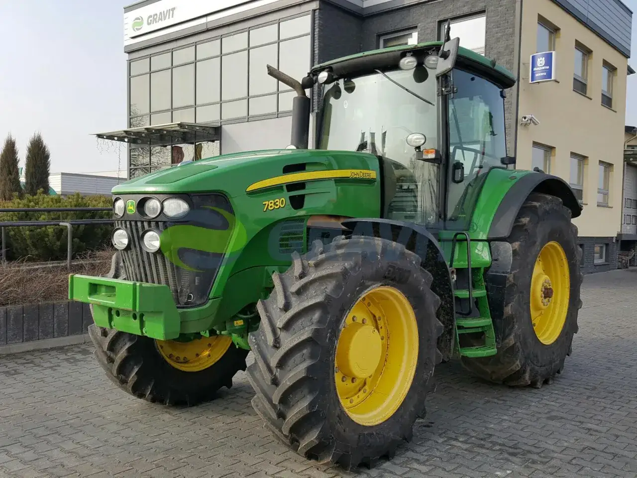 Potężny John Deere 7830, zielony traktor z żółtymi felgami, gotowy do pracy na polu przed nowoczesnym budynkiem.