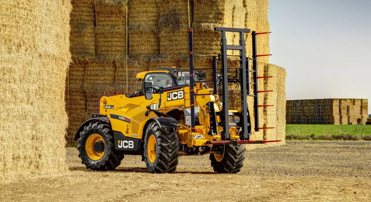 Żółty ładowacz teleskopowy JCB 531-70 z widłami do bel stoi na polu wśród stogów słomy.
