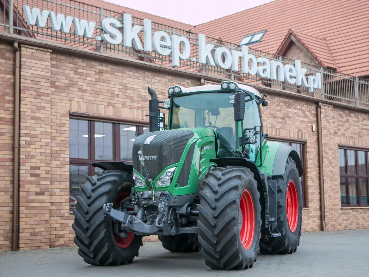Potężny zielony traktor Fendt 936 Vario z czerwonymi felgami przed budynkiem sklepu.