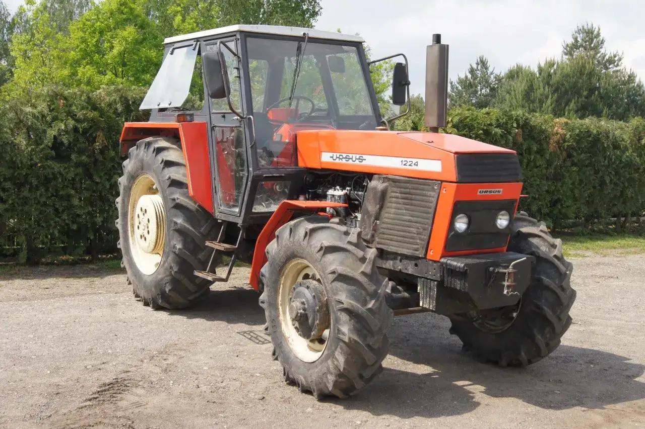 Pomarańczowy Ursus 1224 z kabiną stoi na drodze gruntowej, otoczony zielenią.