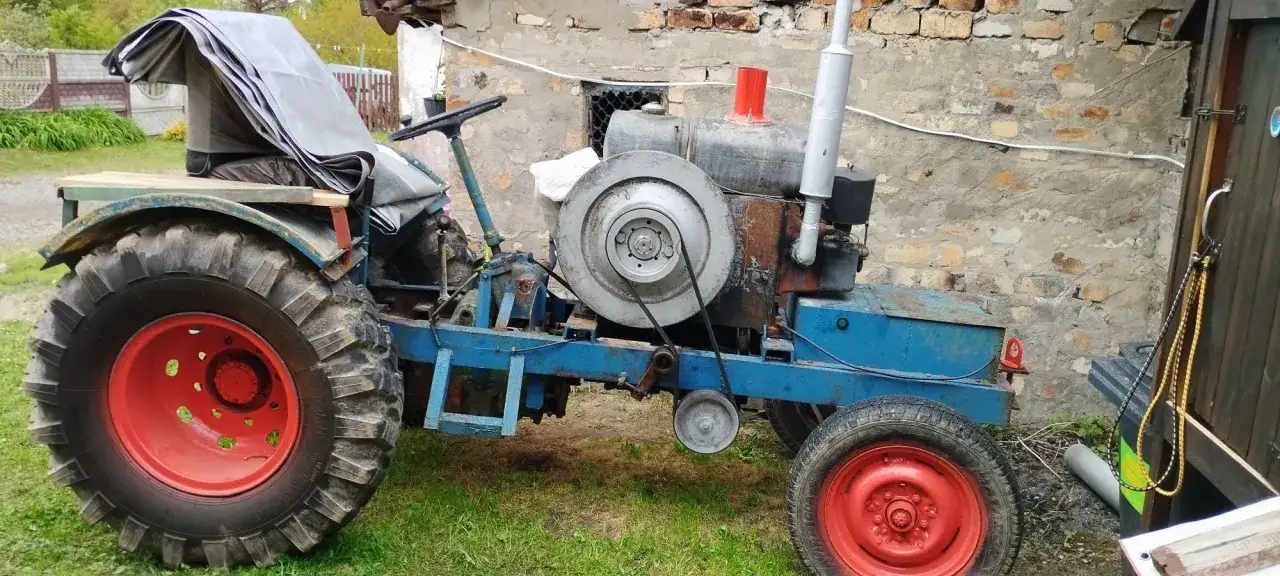 Stary, niebieski traktor, przypominający esioka, z plandeką na siedzeniu, stoi na trawie obok ceglanego budynku.