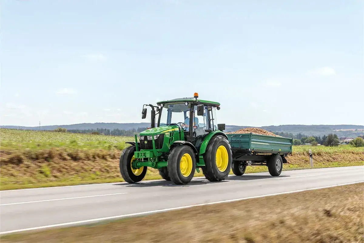 Zielony John Deere 5075e z przyczepą pełną ziemniaków jedzie po drodze wśród pól.